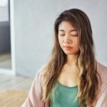 Close up portrait of young Asian woman sitting and meditating an empty room at home. Mental health, mindfullness, wellbeing, balance concept. Minimalistic interior.